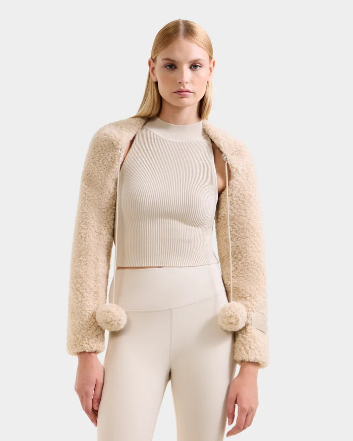 A woman with straight blonde hair wears the Rudsak VERITY - 8525666 Vanilla women’s faux shearling bolero, draped over a cream ribbed top and high-waisted pants, creating a chic cold-weather look against a plain light background.