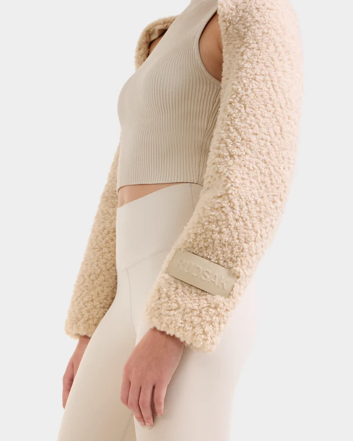 Wearing the VERITY - 8525666 Vanilla faux shearling bolero by Rudsak with a ribbed sleeveless crop top and high-waisted light neutral leggings—an ideal après-ski style set against a plain background.