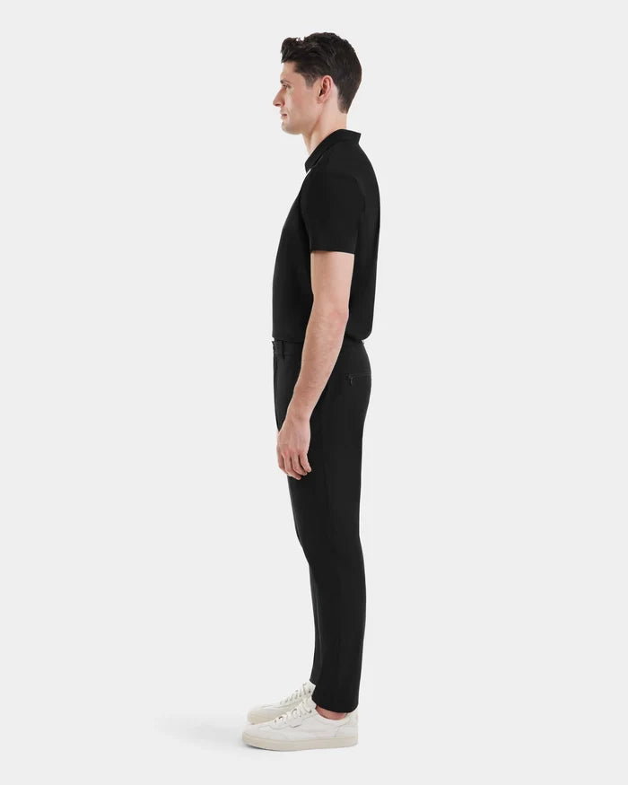 A man stands in profile facing right, wearing the VERNON - 6525025 Black short-sleeve polo shirt by Rudsak, paired with tapered-leg trousers and white sneakers—an effortlessly modern look against a plain white background.