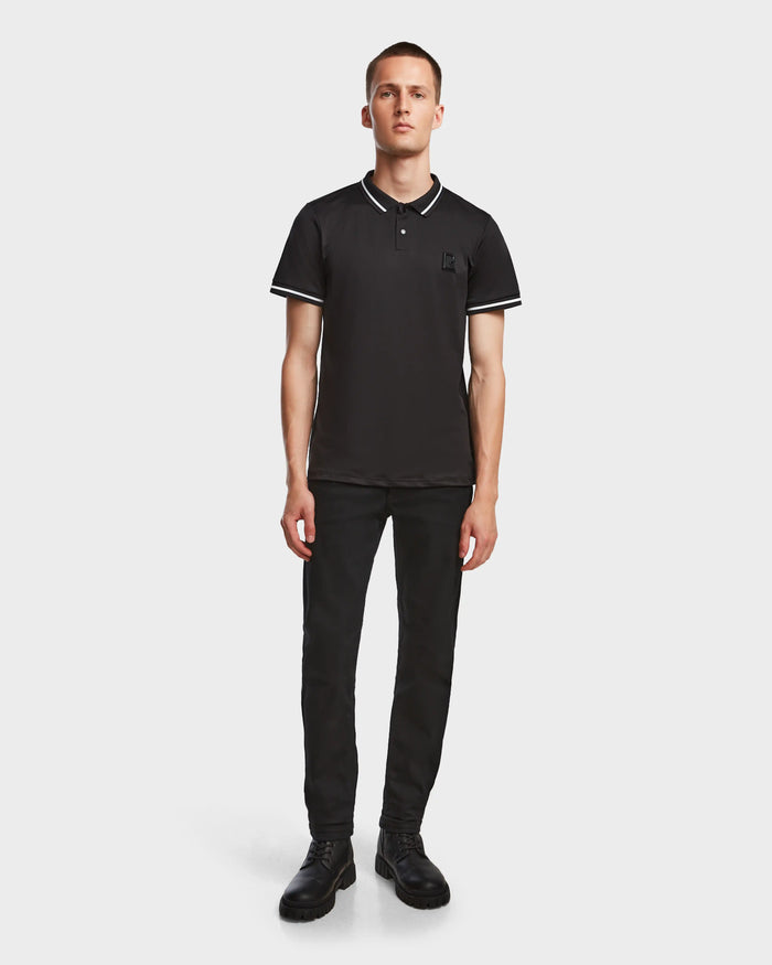 A man faces forward wearing the Rudsak VICK - M523004 Black tailored-fit men’s polo shirt with white trim, paired with black pants and shoes, set against a plain light background.