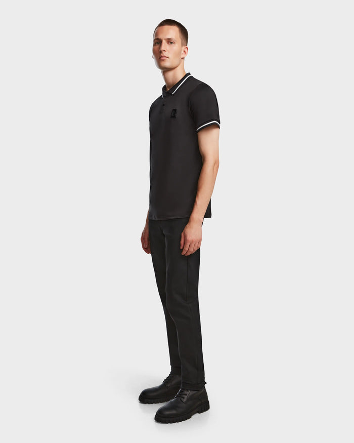 A man stands against a plain background, wearing the Rudsak VICK - M523004 Black tailored fit men’s polo shirt with white trim, paired with black pants and black lace-up boots. He faces slightly to the side with a neutral expression.