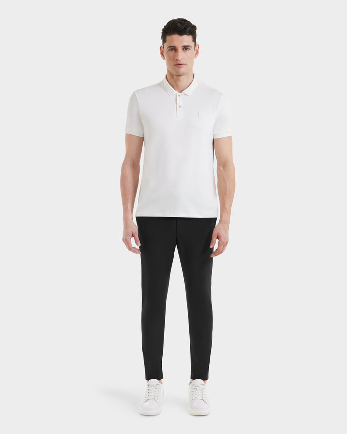 A man stands facing forward against a plain white background, wearing Rudsaks VICK - M523004 Optic White tailored fit men’s polo shirt, black slim-fit pants, and white sneakers.