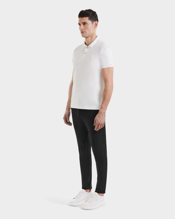 A man stands against a plain background wearing the VICK - M523004 Optic White men’s polo shirt by Rudsak, paired with black slim-fit pants and white sneakers, facing forward with his arms relaxed at his sides.
