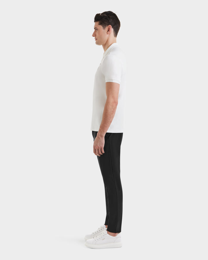 A man in profile faces left, wearing the Rudsak VICK - M523004 Optic White men’s polo shirt, paired with black pants and white sneakers, against a plain white background.