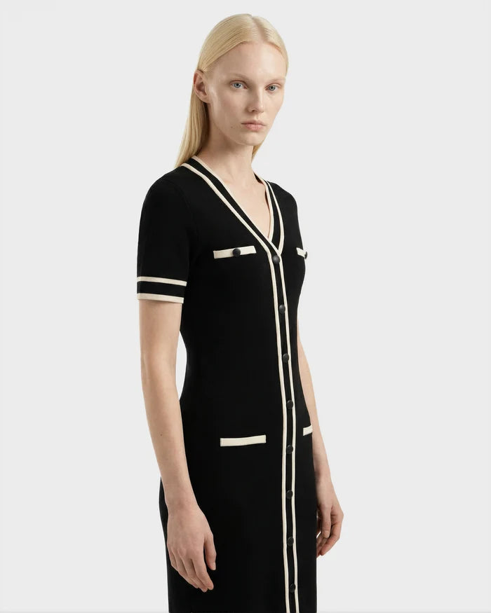 A woman with straight blonde hair wears the Rudsak VINNIE - 8526038 Black: a fitted black knit midi dress with short sleeves, white trim, and white-accented pockets, styled for everyday wear against a plain light background.