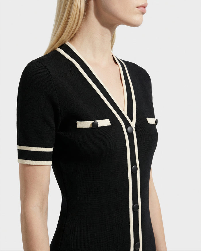A woman with straight blonde hair models the Rudsak VINNIE - 8526038 Black dress, featuring a V-neckline, white trim, front buttons, and decorative pocket flaps. She is pictured from shoulders to waist, facing sideways.