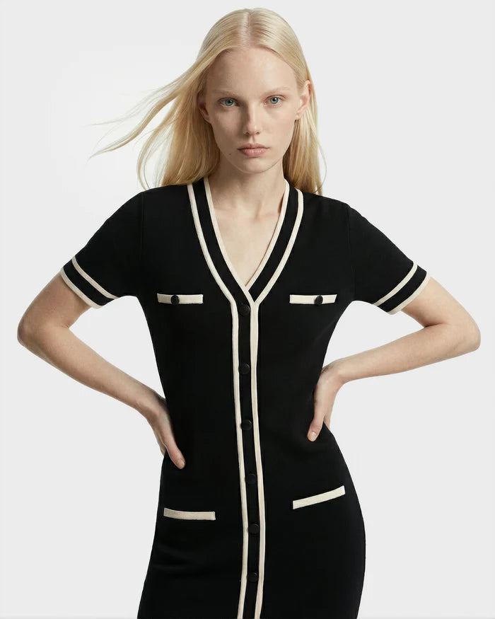 A woman with long blonde hair poses against a white background, wearing the Rudsak VINNIE - 8526038 Black dress featuring a V-neckline, white trim, and front buttons, with her hands on her hips.