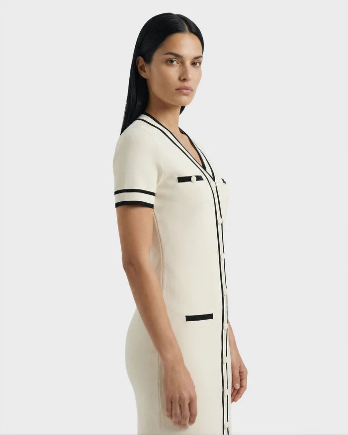 A woman with long dark hair stands sideways against a plain light background, wearing the Rudsak VINNIE - 8526038 Vanilla fitted knit midi dress in cream with black trim and pocket details.