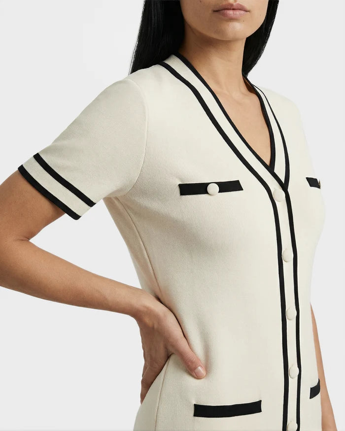 A woman stands confidently with one hand on her hip, wearing the Rudsak VINNIE - 8526038 Vanilla short-sleeved cream cardigan with black trim over a V-neck dress, set against a plain background.