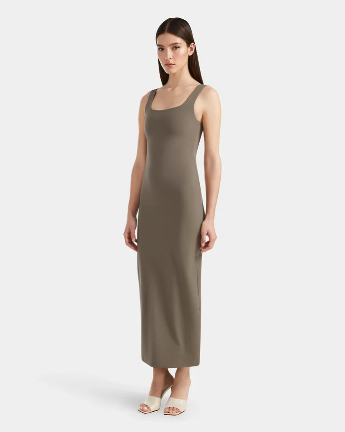 A woman wearing the Rudsak WANDA - 8524035 Cement sleeveless, ankle-length taupe maxi dress made from stretch performance fabric and white open-toe heels stands against a plain white background.