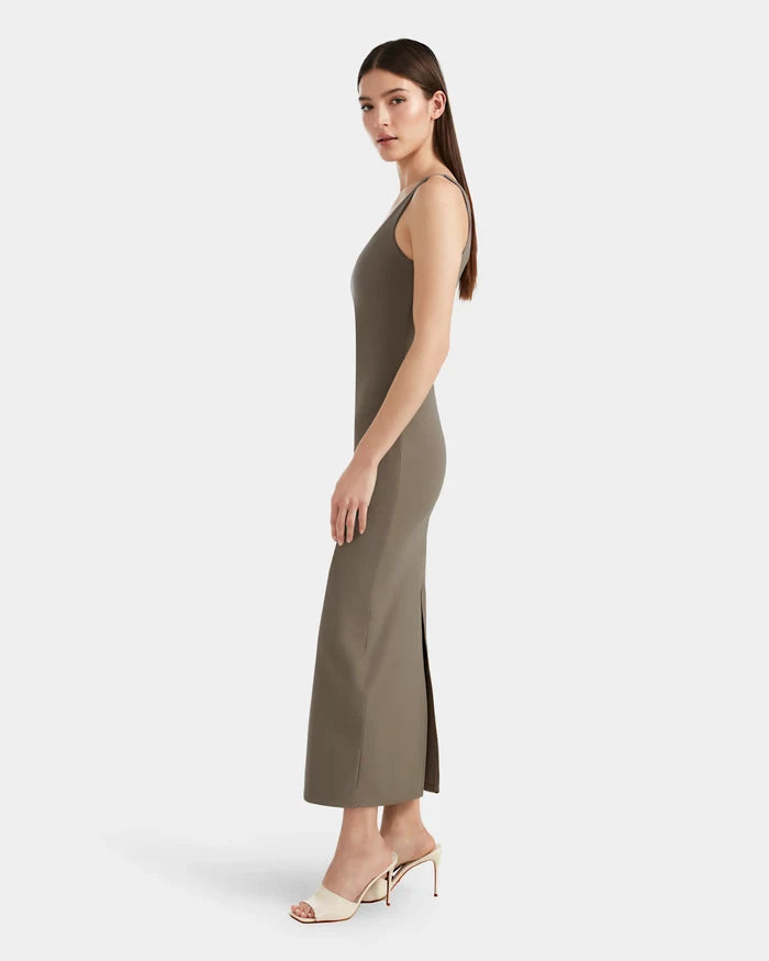 A woman in profile wears the Rudsak WANDA - 8524035 Cement sleeveless, fitted maxi dress in taupe stretch fabric with a side slit. With long straight hair and white heeled sandals, she poses elegantly against a plain white background.