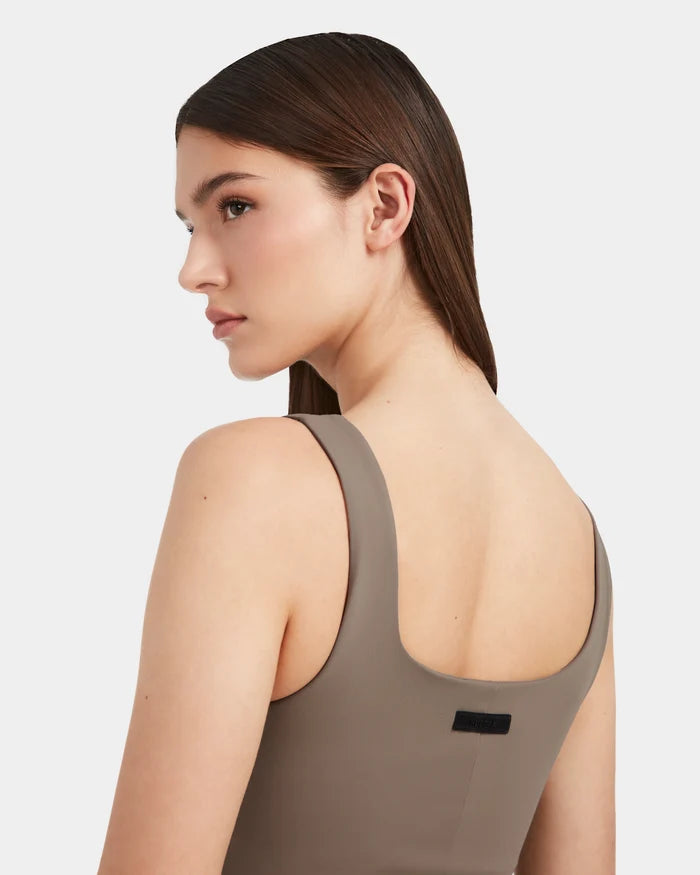 A woman with straight brown hair poses in the WANDA - 8524035 Cement sleeveless taupe top by Rudsak, featuring a contemporary scoop back, set against a plain light background.