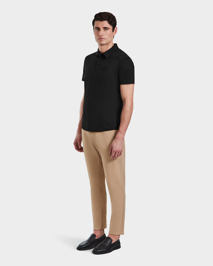 A man stands against a white background wearing Rudsaks WELLS - 6525017 Black tailored-fit short-sleeve polo, beige slim-fit pants, and black loafers—a modern essential look.