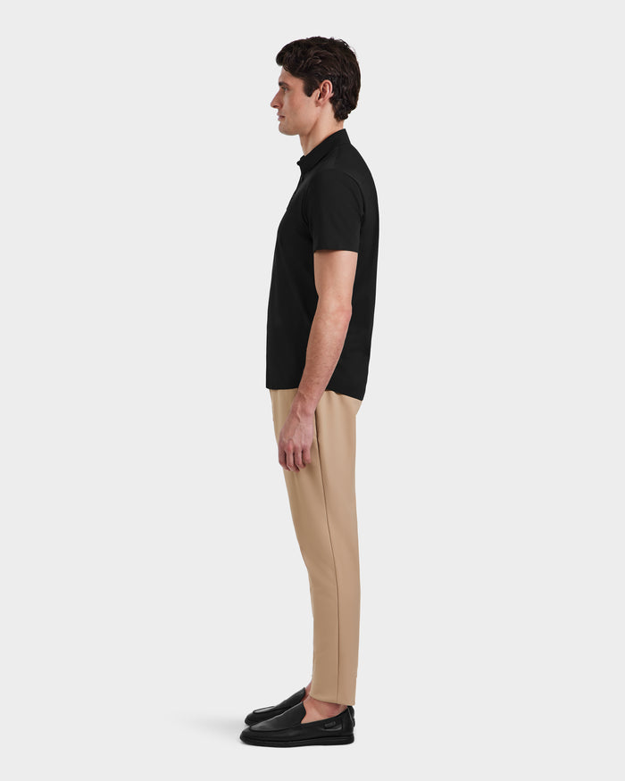 A man stands in profile facing left, wearing the Rudsak WELLS - 6525017 Black polo crafted from lightweight technical fabric, paired with tailored beige pants and black loafers against a plain white background.