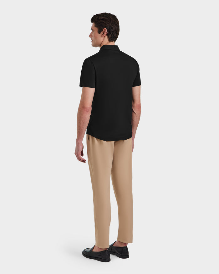 A man faces away, wearing the Rudsak WELLS - 6525017 Black short-sleeve polo made from lightweight technical fabric, paired with beige pants and black shoes against a plain white background—a modern essential with a tailored fit.