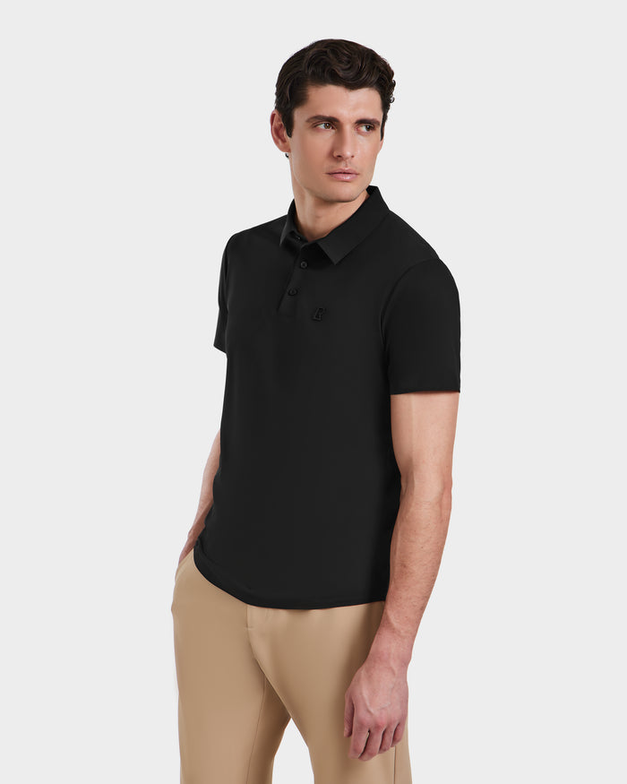 A man wearing the Rudsak WELLS - 6525017 Black tailored-fit short-sleeve polo with beige pants stands against a light background, looking right with one hand in his pocket—an effortless modern essential.