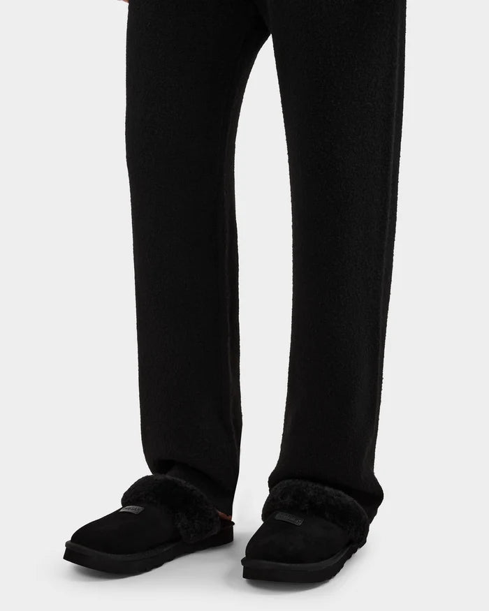 A person wears loose black pants and Rudsak’s WILBER - 6225509 Black faux shearling slippers, standing on a white background. The focus is on their lower legs and feet, showcasing the cozy men’s style by Rudsak.