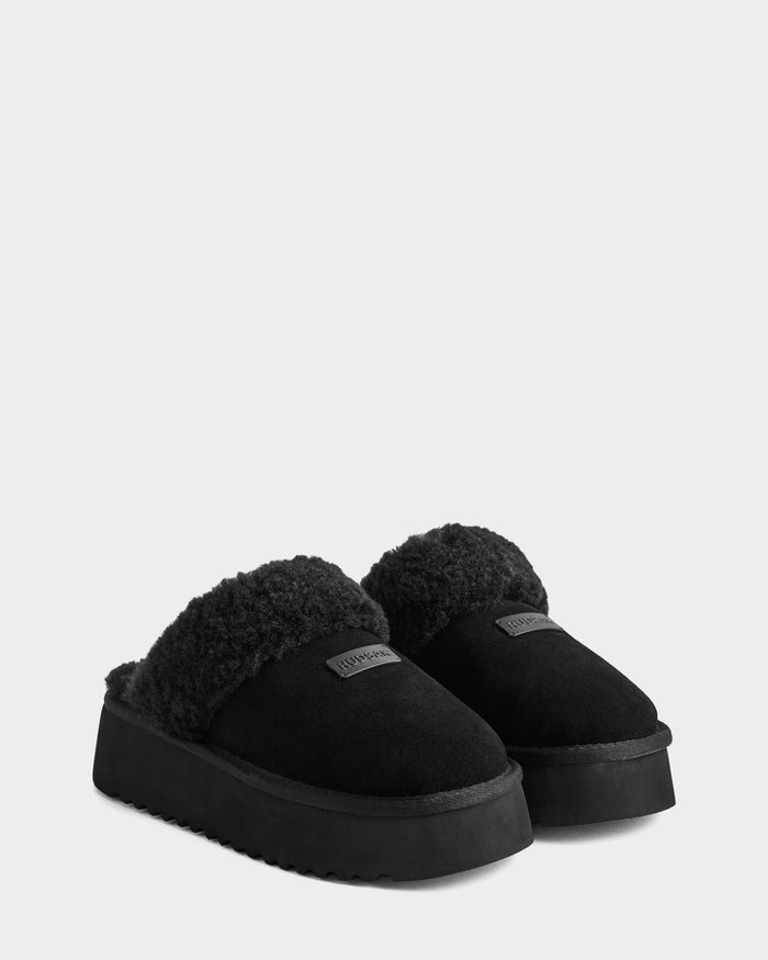 The Rudsak WILMA - 8225582 Black women’s slippers feature a suede-like black finish, thick soles, plush faux shearling lining, and a label on the upper front, shown against a plain white background.