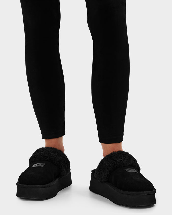 A person in black leggings wears Rudsak’s WILMA - 8225582 Black women’s slippers featuring faux shearling and thick soles, standing on a light gray background.