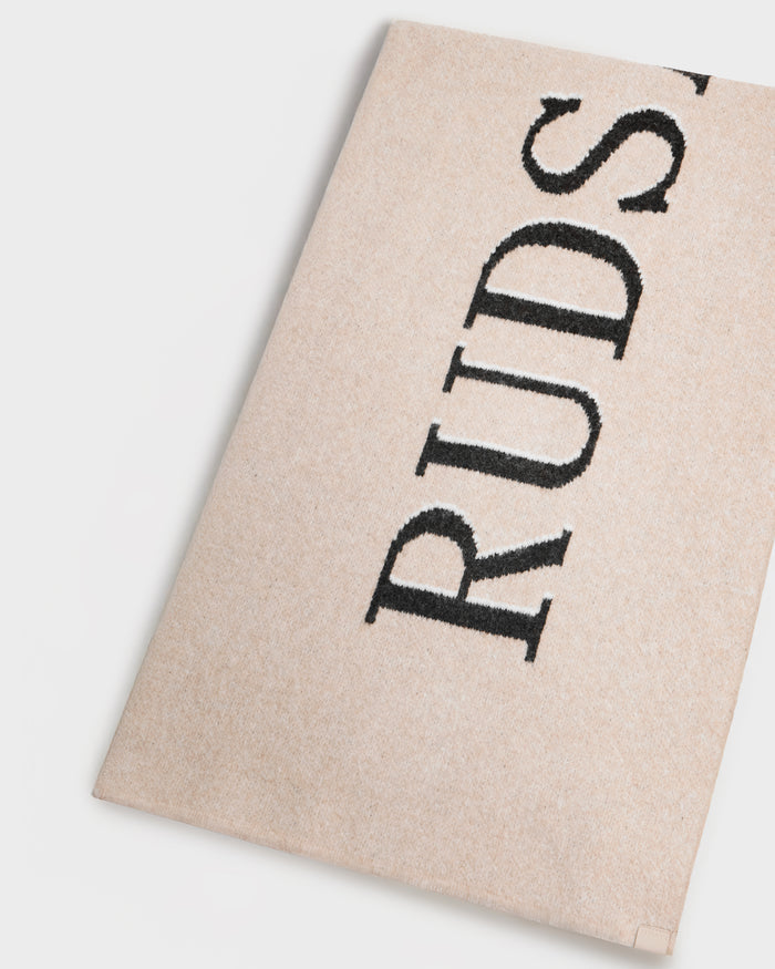 A beige textured surface shows partial vertical black "RUDS" text, photographed on white. It’s the WYATT - 6425039 Vanilla scarf by Rudsak—a men’s winter essential combining style and warmth.