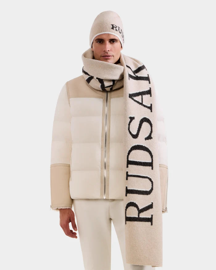 A person wears the Rudsak WYATT - 6425039 Vanilla puffer jacket with matching pants, a beige beanie, and a bold RUDSAK scarf, highlighting standout winter style against a plain white background.