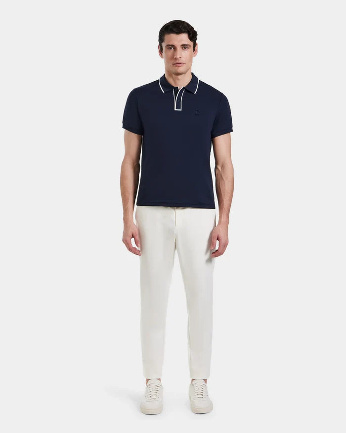 A man faces forward wearing the Rudsak XAVIER - 6524027 Navy polo shirt with white piping, paired with white pants and sneakers, against a plain white background.