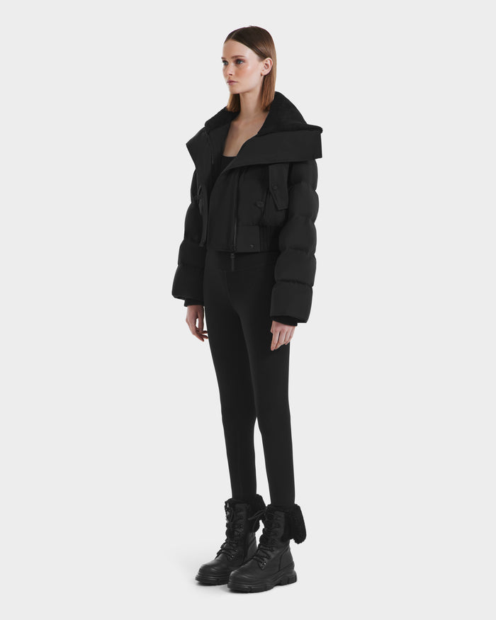 A woman stands against a plain background, wearing the Rudsak YVETTE - L125557 Black oversized puffer jacket with RDS-certified down, paired with black leggings and combat boots. She faces slightly to the side and maintains a neutral expression.