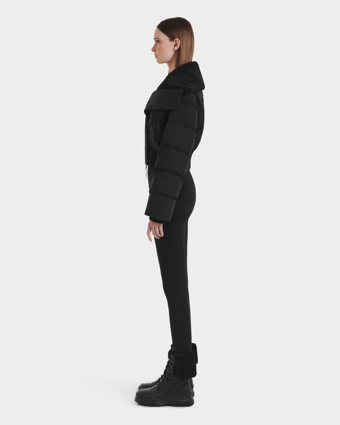 A woman in profile stands against a plain background, wearing Rudsak's YVETTE - L125557 Black women’s cropped down jacket with an oversized collar, paired with black leggings and black fur-trimmed lace-up boots.