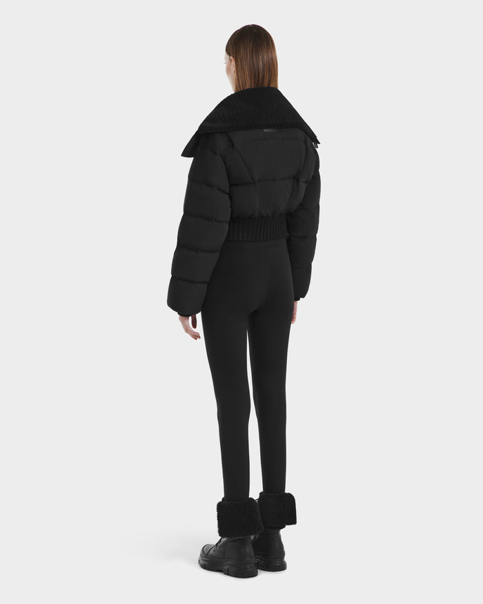 A person with straight brown hair faces away, wearing the Rudsak YVETTE - L125557 Black women’s cropped down jacket with an oversized collar, paired with black leggings and black boots with fuzzy cuffs against a plain white background.