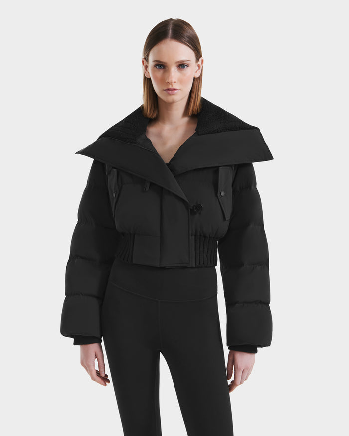 A woman with straight brown hair models the Rudsak YVETTE - L125557 Black cropped down jacket with an oversized collar and high-waisted black leggings against a plain white background.
