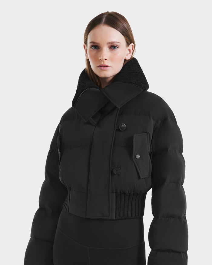 A woman with straight brown hair models the Rudsak YVETTE - L125557 Black cropped down jacket with RDS-certified down, high oversized collar, and front buttons, paired with black fitted pants against a plain light background.