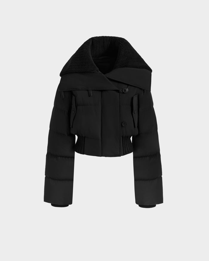 The Rudsak YVETTE - L125557 Black is a women’s cropped down jacket with an oversized collar, long sleeves, large buttons, and a slightly cinched waist, shown on a plain light gray background.