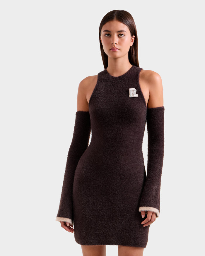 A young woman models the Rudsak ZARAH - 8525696 Espresso dress, a fitted brown knit with detached matching sleeves and a white "R" patch, standing against a plain light gray background.