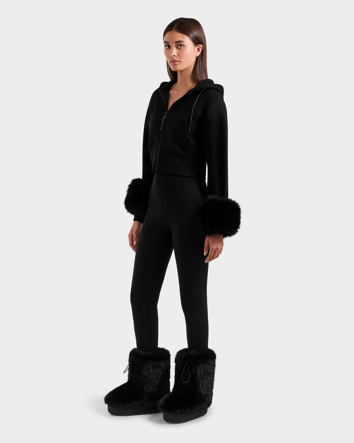A woman with straight dark hair and a neutral expression stands against a plain background wearing the Rudsak ZILLIA - 8525673 Black women’s cropped hoodie with faux fur cuffs, matching black leggings, and black furry boots.