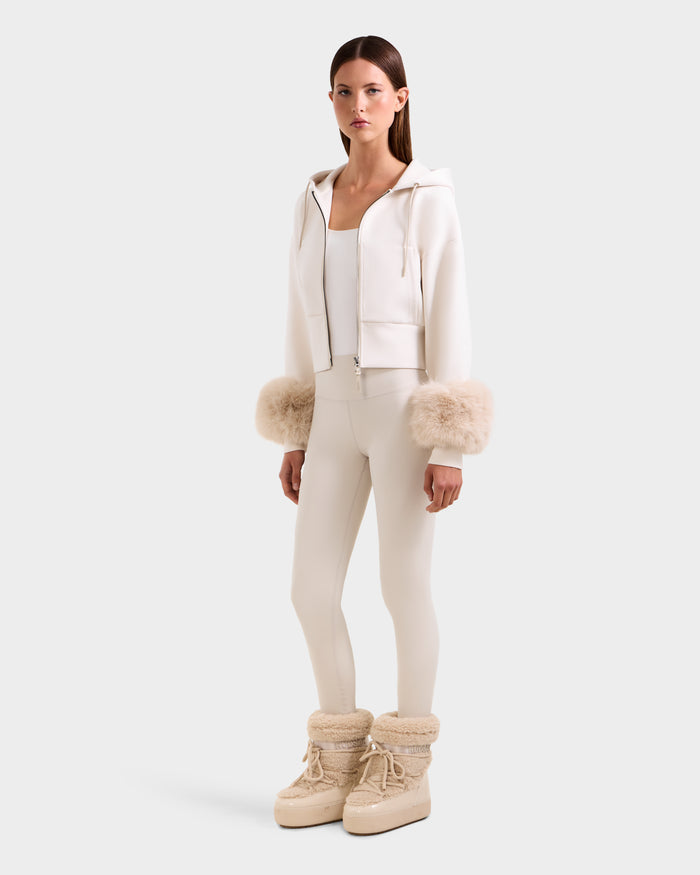 A woman poses against a plain background wearing the Rudsak ZILLIA - 8525673 Vanilla: a chic all-cream outfit featuring a cropped zip-up hoodie with faux fur cuffs, matching fitted leggings, a tank top, and beige winter boots with fur trim.
