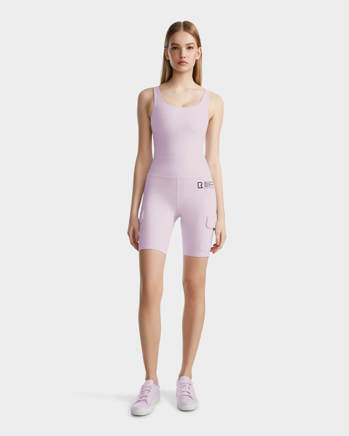 A young woman faces forward, wearing the Rudsak ZORA - 8524066 Lilac Foam fitted sleeveless bodysuit with matching high-performance biker shorts and lavender sneakers, set against a plain white background.