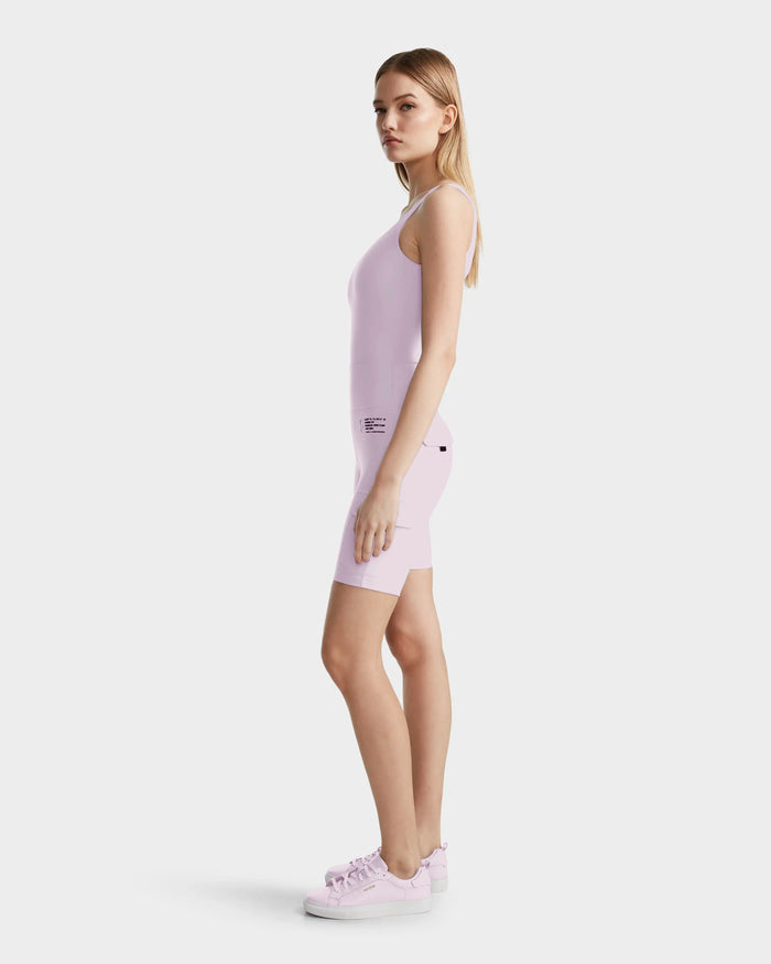 A woman stands sideways wearing the Rudsak ZORA - 8524066 Lilac Foam sleeveless fitted romper, crafted from high-performance stretch fabric, paired with matching pink sneakers against a plain white background.