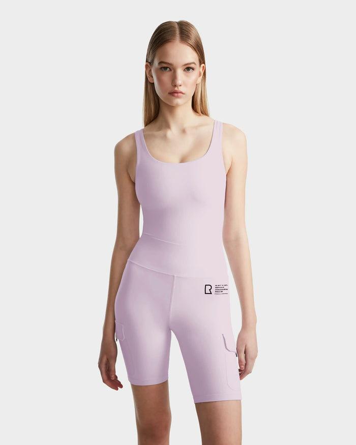 A young woman stands facing forward in the Rudsak ZORA - 8524066 Lilac Foam romper, a fitted, sleeveless short piece with small black text on the left thigh, made from high-performance stretch fabric. The background is plain white.