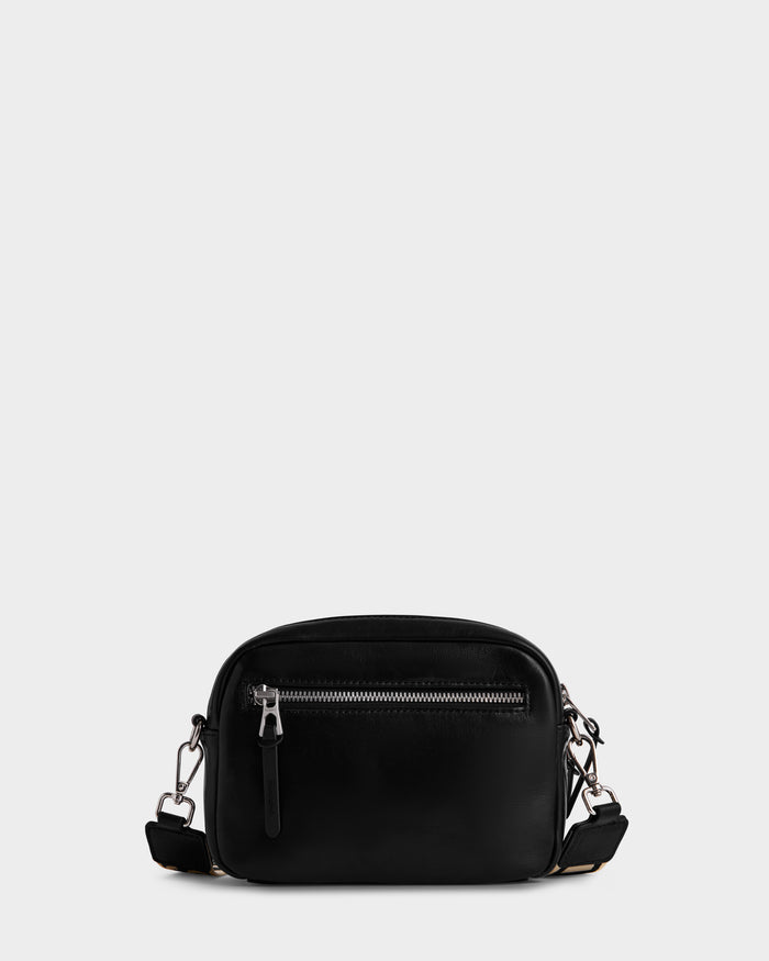 The Rudsak ZOYA L - 8325543 Black is a small black crossbody bag in smooth leather, with a front zip pocket, silver hardware, and an adjustable logo strap, shown against a plain light gray background.