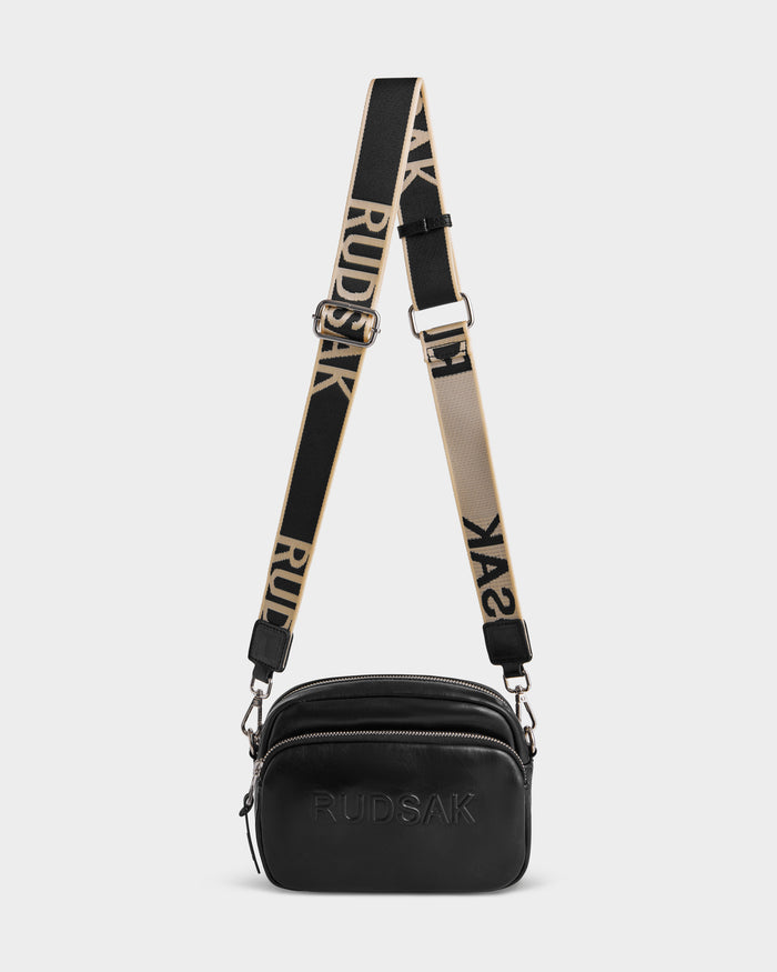 The ZOYA L - 8325543 Black by Rudsak is a compact black leather crossbody with embossed branding, two zippered compartments, and a wide adjustable beige-and-black logo strap.